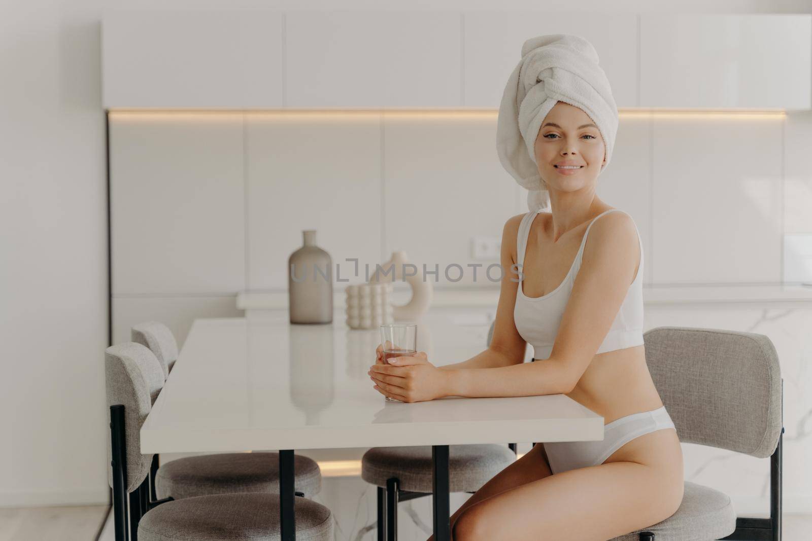 First thing in the morning. Relaxed slender woman sitting in stylish kitchen after taking shower with glass of pure mineral water, wears white underwear and towel over wet hair. Weightloss and dieting