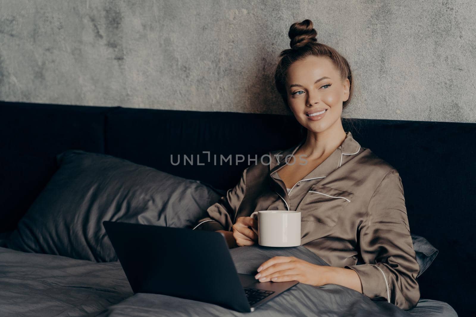 Lazy relaxed morning time. Smiling beautiful young female lying in bed in satin pajama with cup of coffee in her hand while checking last news on internet online on laptop, enjoying free time at home