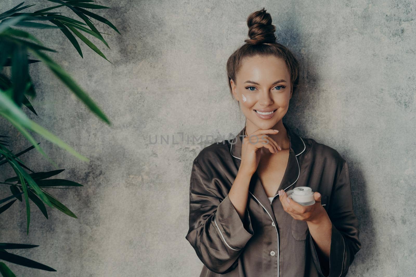 Happy female wear silk brown pajama with moisturizing cream in her hand during morning beauty routine by vkstock