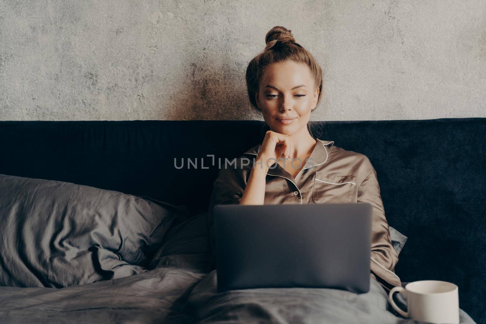 Positive relaxed young woman surfing internet for favorite film to watch on laptop while spending lazy day off at home, lying in bed at home and looking at computer monitor, dressed in pajama