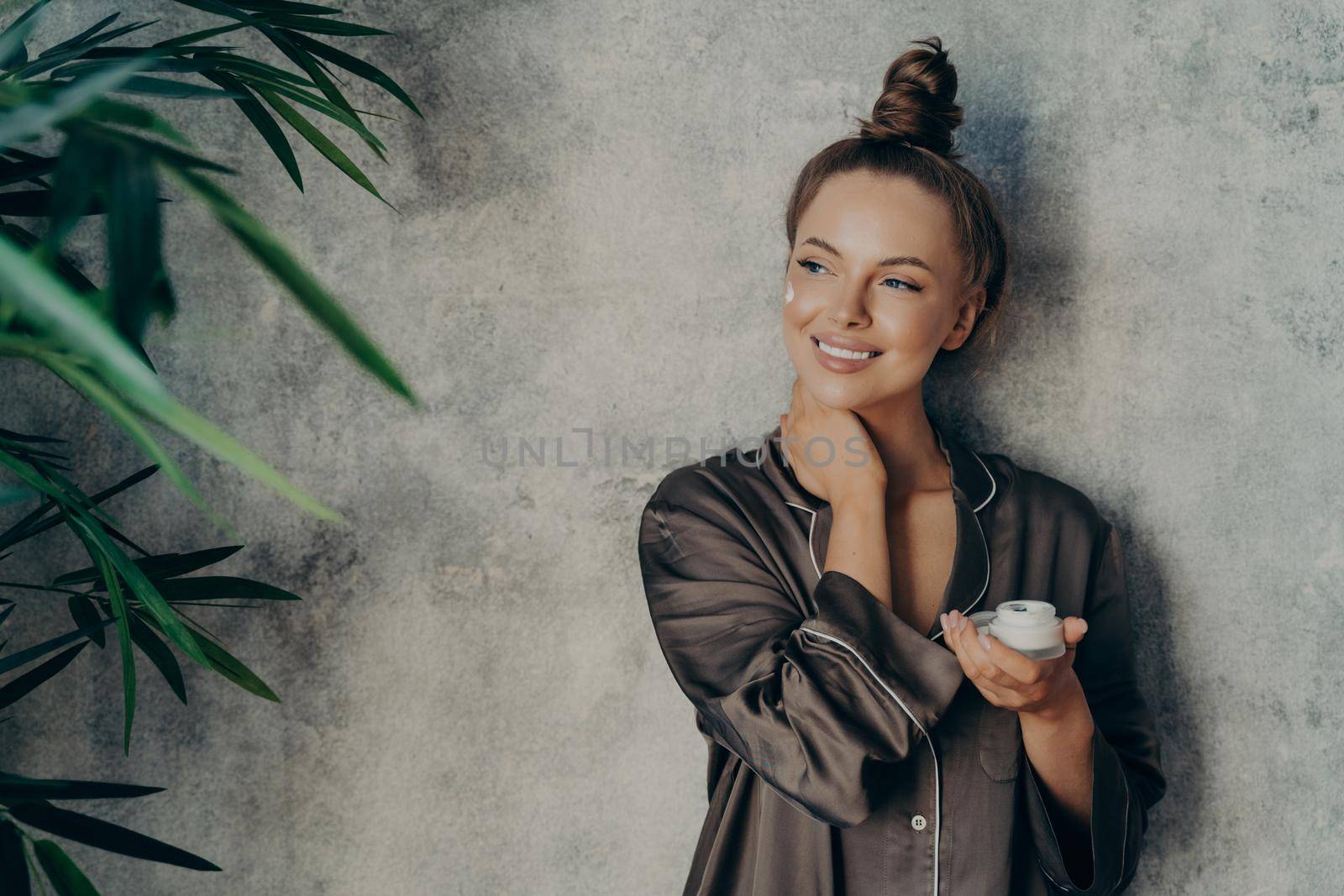 Young beautiful woman with glowing healthy skin broadly smiling while holding face cream by vkstock
