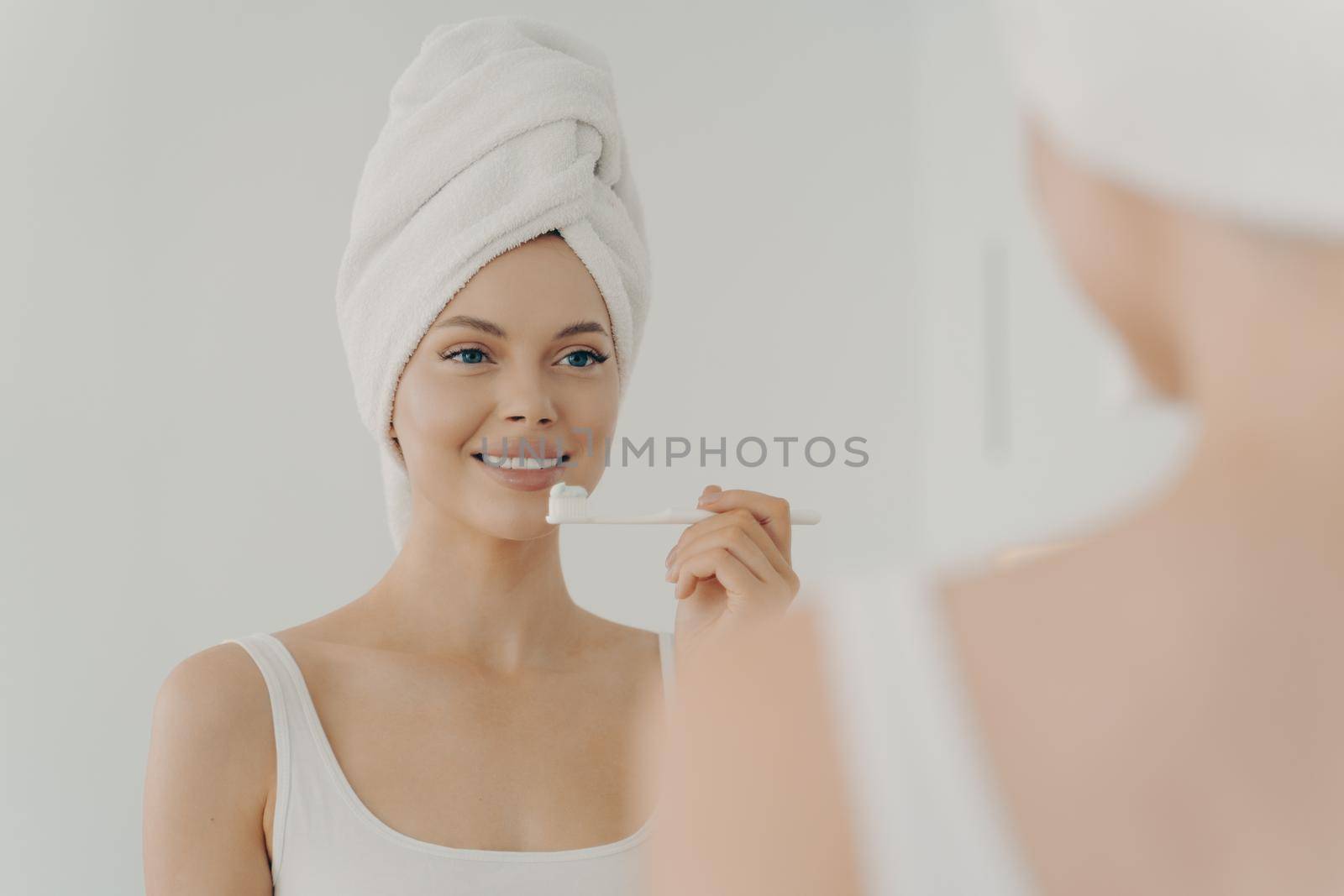 Healthy beautiful woman with towel on head after shower having toothy smile while gently brushing her teeth by vkstock