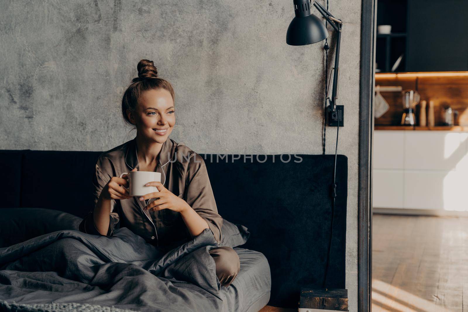 Happy smiling caucasian female in pajama enjoying morning cup of coffee while sitting in bed after waking up before starting her busy work day, relaxing in bedroom of contemporary apartment or hotel