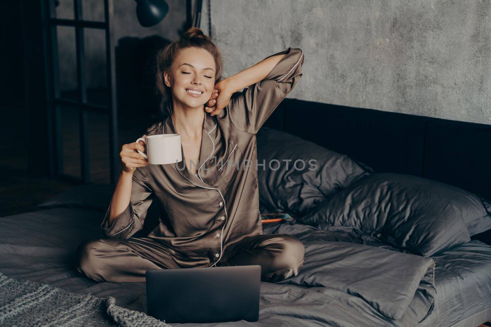 Relaxed female freelancer having cup of coffee stright after waking up in bed while checking new messages and emails on laptop, dressed in pajama. Woman with closed eyes ready to start remote work