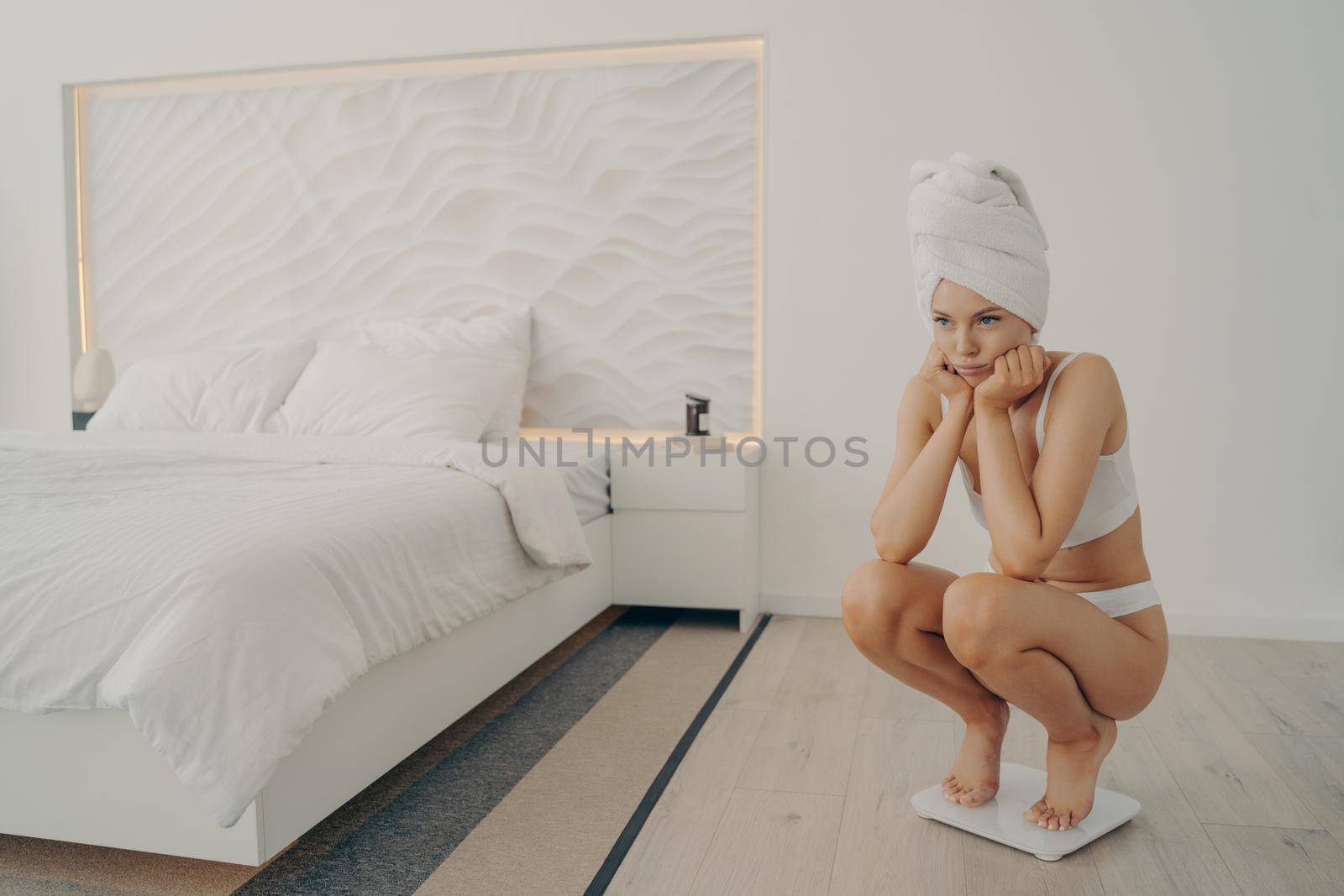 Young sad beautiful woman in white underwear standing crouched on electronic smart scales in stylish bedroom background, unhappy with what she saw. Weight loss and dieting concept