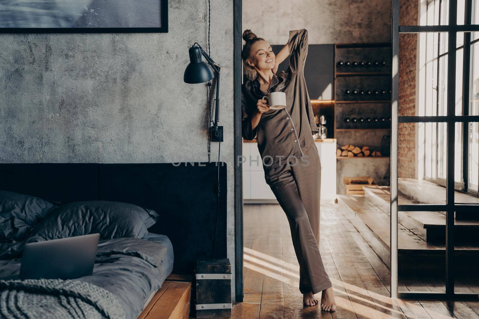 Attractive slim caucasian young woman in satin pajama with cup of coffee in her hand stretching in bedroom by vkstock