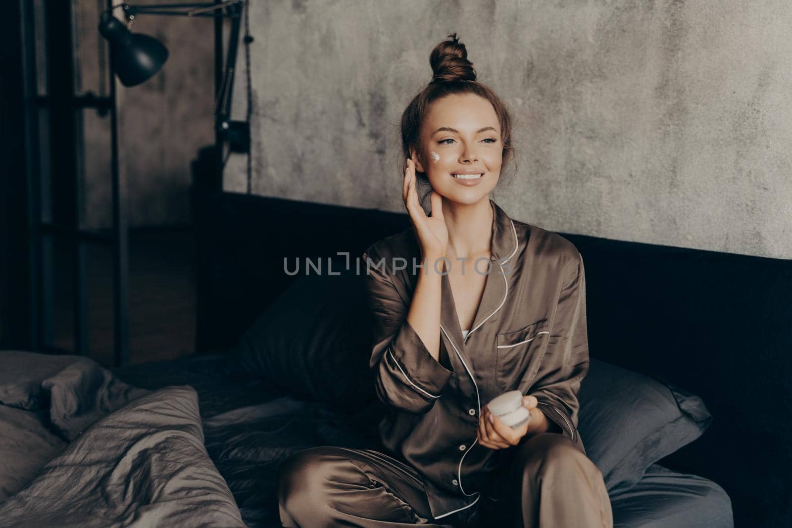 Young pretty european woman in silk pajama takes care of skin and body after waking up in morning at home while applying moisturizing face cream, getting ready for work. Beauty and skincare concept