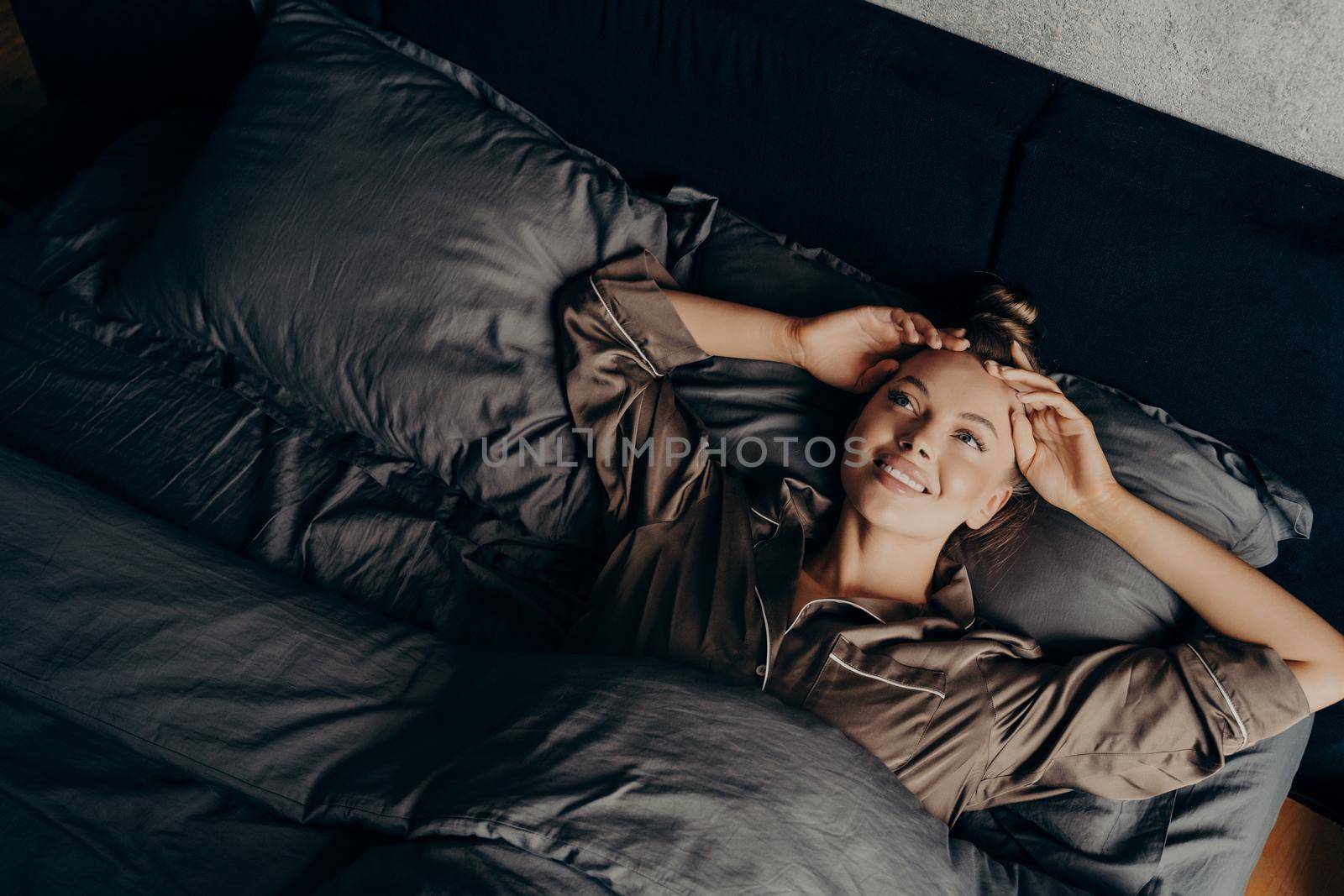 Beautiful young girl in satin comfy pajama waking up while lying in bed at home by vkstock