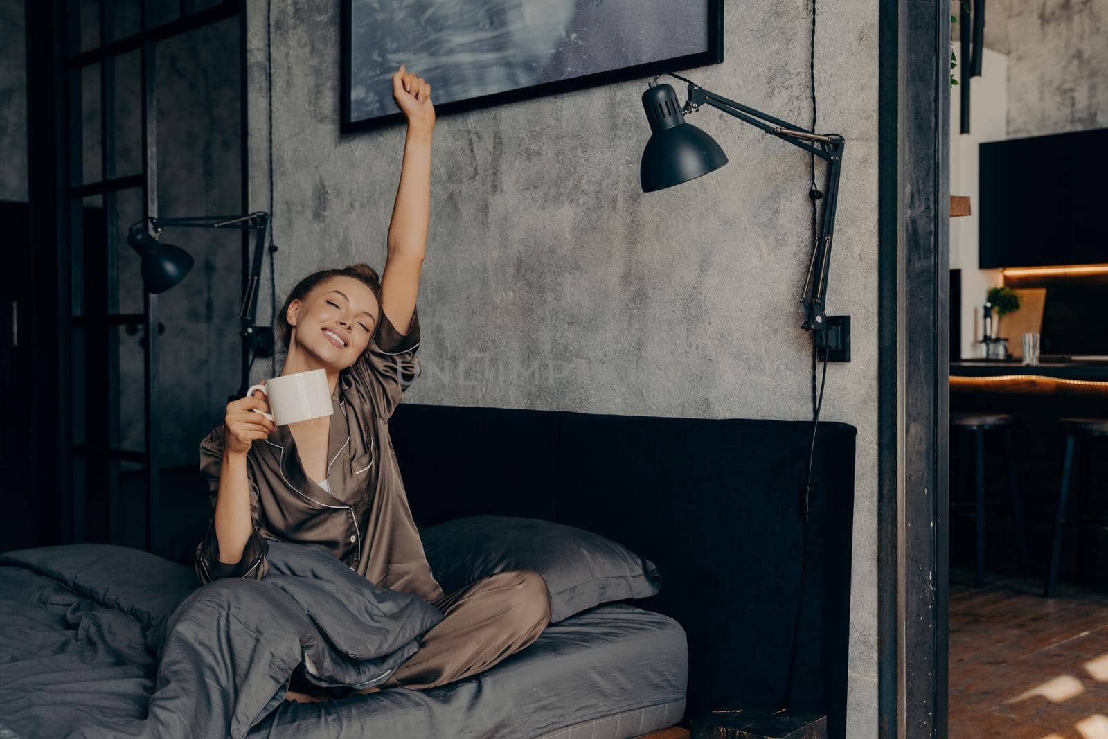 Young beautiful woman with closed eyes and hair tied up in bun starting morning with cup of hot coffee, stretching in bed at home while sitting in satin pajama after waking up from night sleep