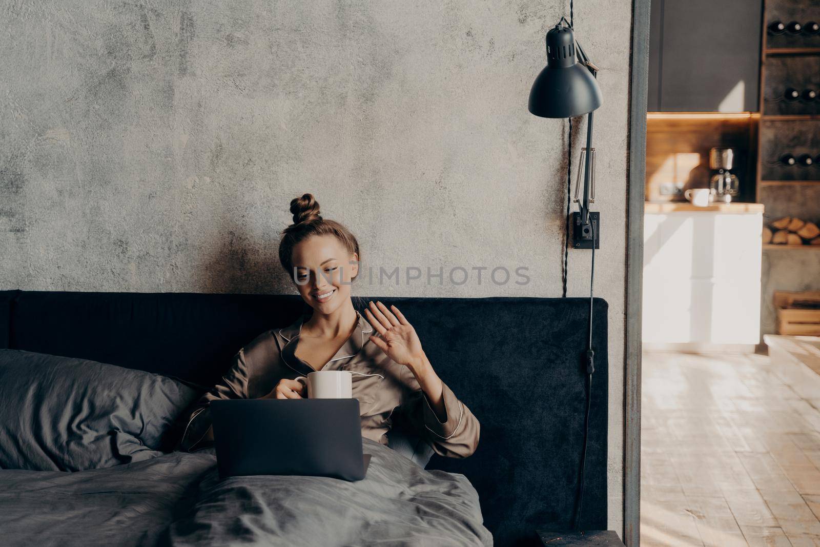 Relaxed calm young woman lying down on bed at home with morning cup of coffee while chatting online with her friends on laptop, smiling and waving to them with hand, dressed in cozy satin pajama