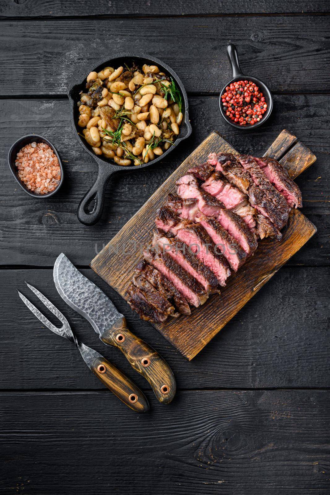 Sliced grilled meat steak Rib eye medium rare, on wooden serving board, with white beans and rosemary in cast iron pan, with meat knife and fork, on black wooden table background, top view flat lay, with copy space for text by Ilianesolenyi