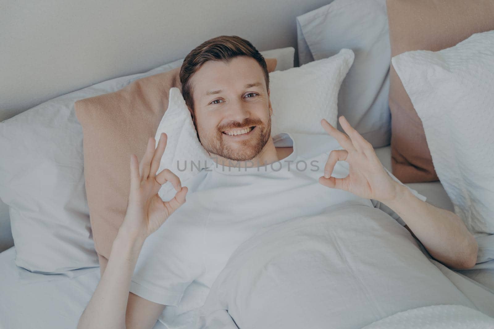Relaxed young bearded man lying in his bed after waking up in early morning, enjoying leisure time while staying at home on weekend, satisfied guy showing ok sign with both hands and looking at camera