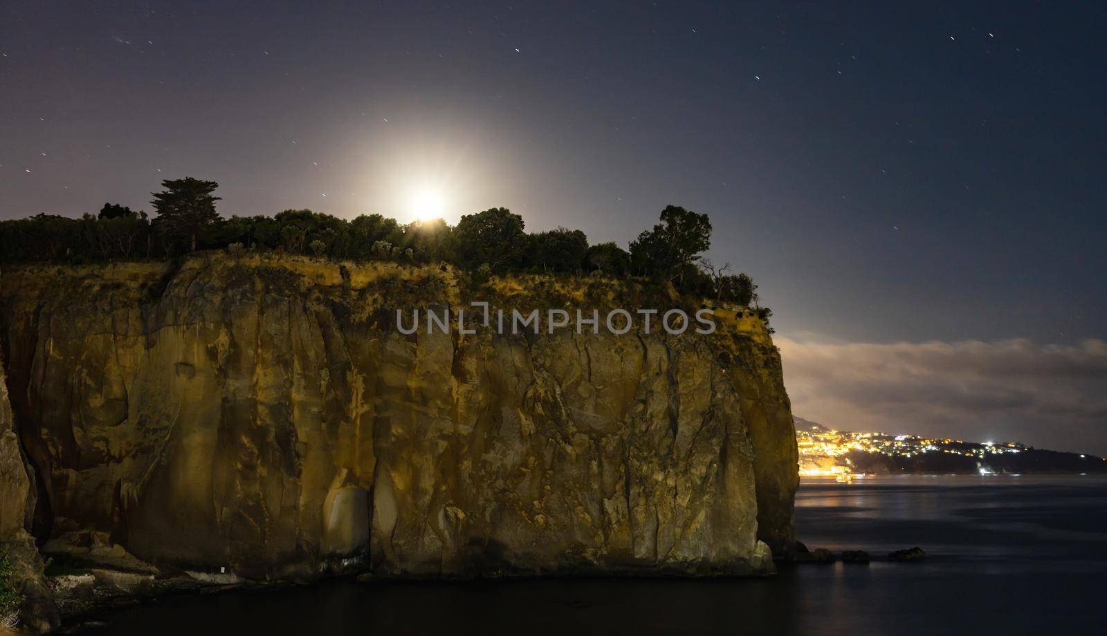 Night landscape of high cliff with glowing moon over it and edge of Sorrento behind