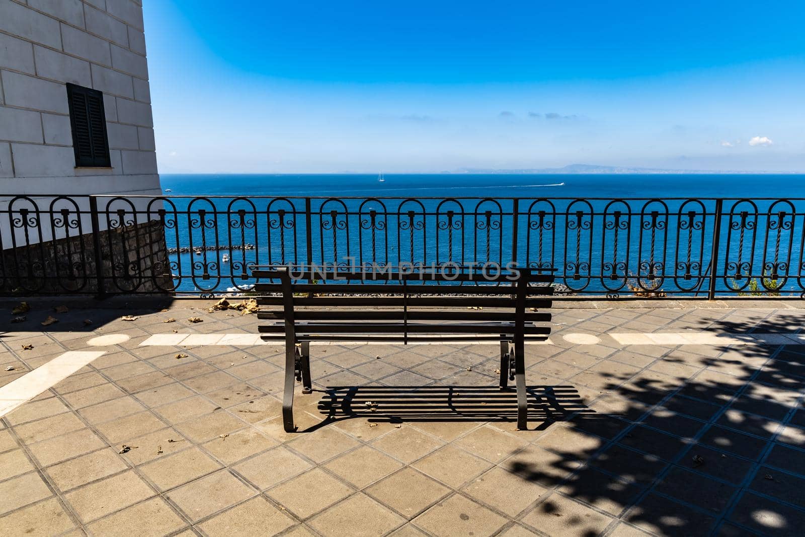 Small bench standing on the edge in front of metal railing with beautiful view to the sea by Wierzchu