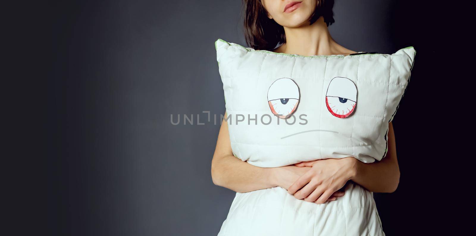 Young girl holds a pillow, on which a displeased face and tired eyes are drawn. Creative idea about sleep problems, insomnia and the importance of quality orthopedic materials.