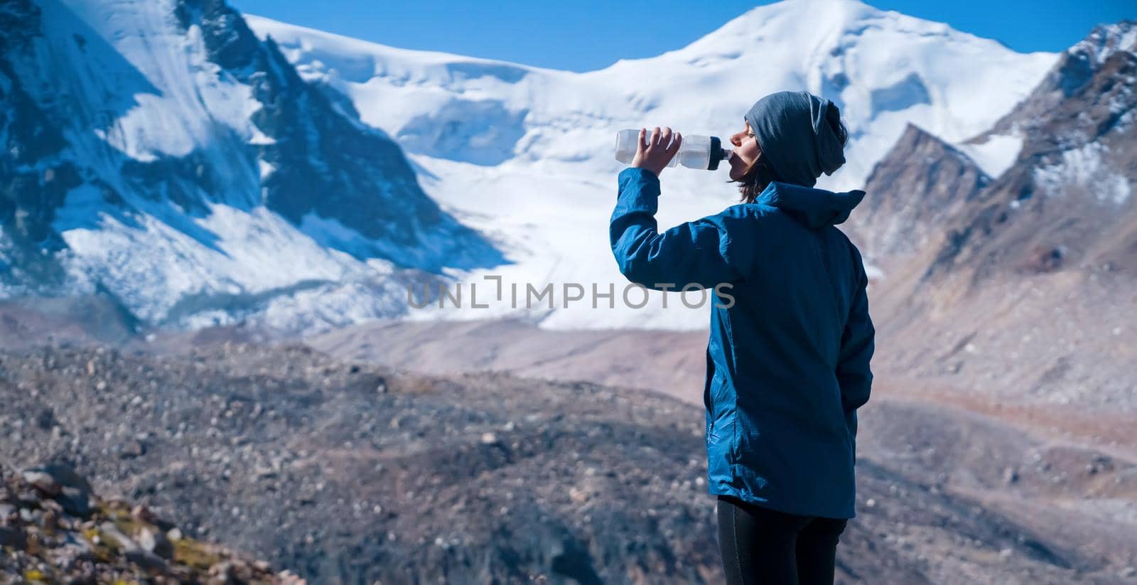Young sports girl stands on the backdrop of a stunning view with snow-capped mountains and drinks clear water from a bottle. Woman is trail running, training, climbing and drinking isotonic.