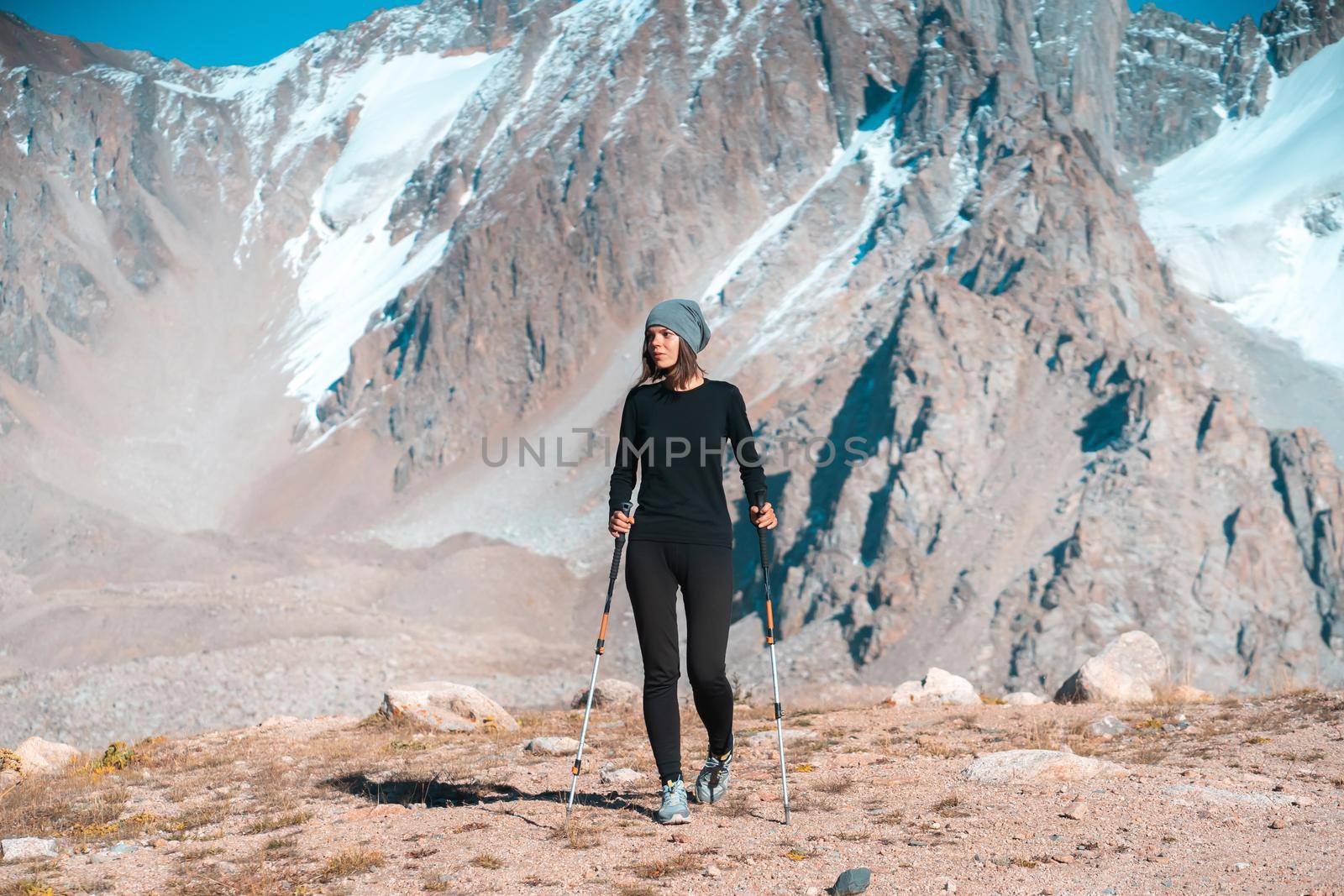 A young beautiful sports girl in thermal underwear with trekking poles walks along the trail among the snow-capped high peaks of the mountains, the traveler walks and climbs tin the national park.