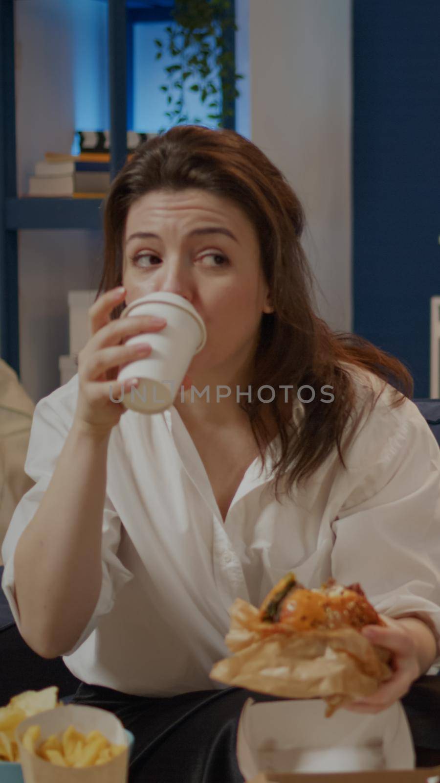 Caucasian woman taking bite from burger and sip of soda sitting on couch at TV. Person eating takeaway food and drinking juice watching television. Adult having unhealthy dinner at home