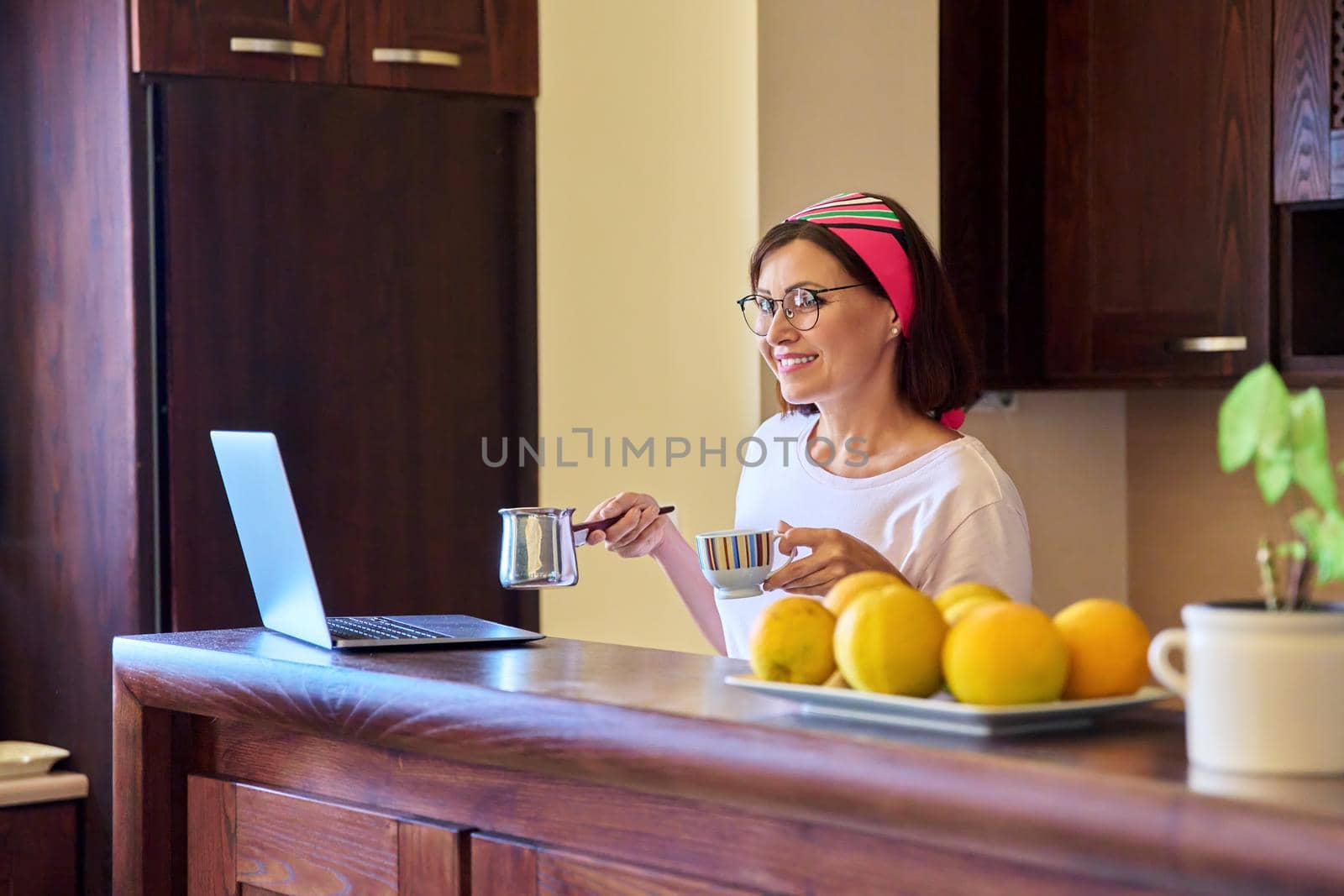 Smiling relaxing woman with morning coffee with cezve turk and laptop in the kitchen at home. Middle-aged female enjoying a drink, technology, lifestyle, home