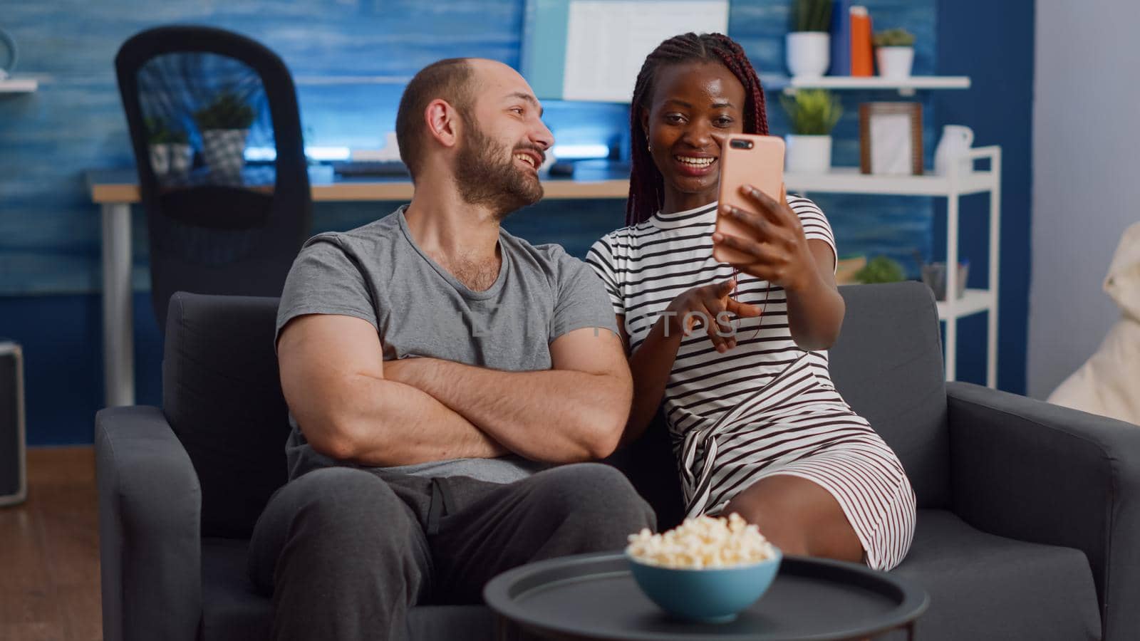 Modern interracial couple talking on video call conference at home. Young multi ethnic partners using internet for remote communication, sitting together on couch in living room.