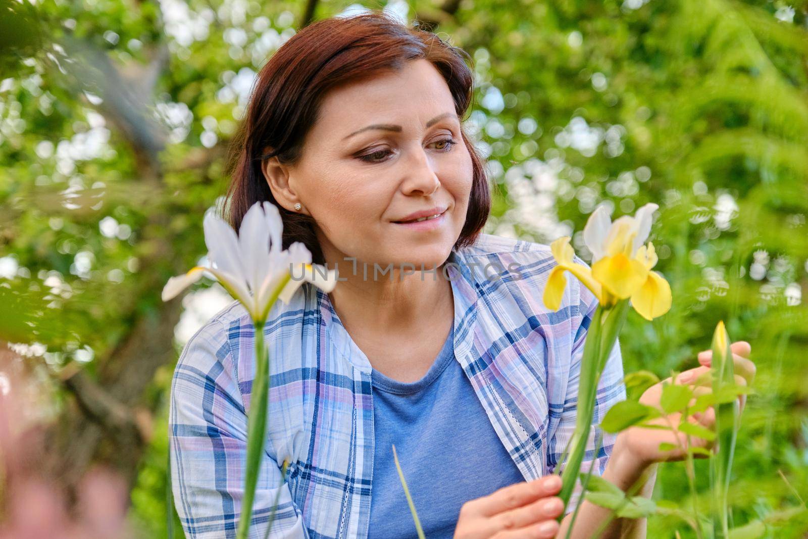 Portrait of 40s woman enjoying iris flowers in a spring garden on a flower bed. Beauty, nature, people of mature age concept