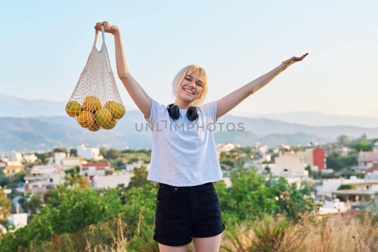 Happy female teenager with eco-friendly mesh bag of farm organic oranges, sunset village mountains. Food trends, buying natural farm fruits, using reusable non-plastic shoppers, fashion and ecology