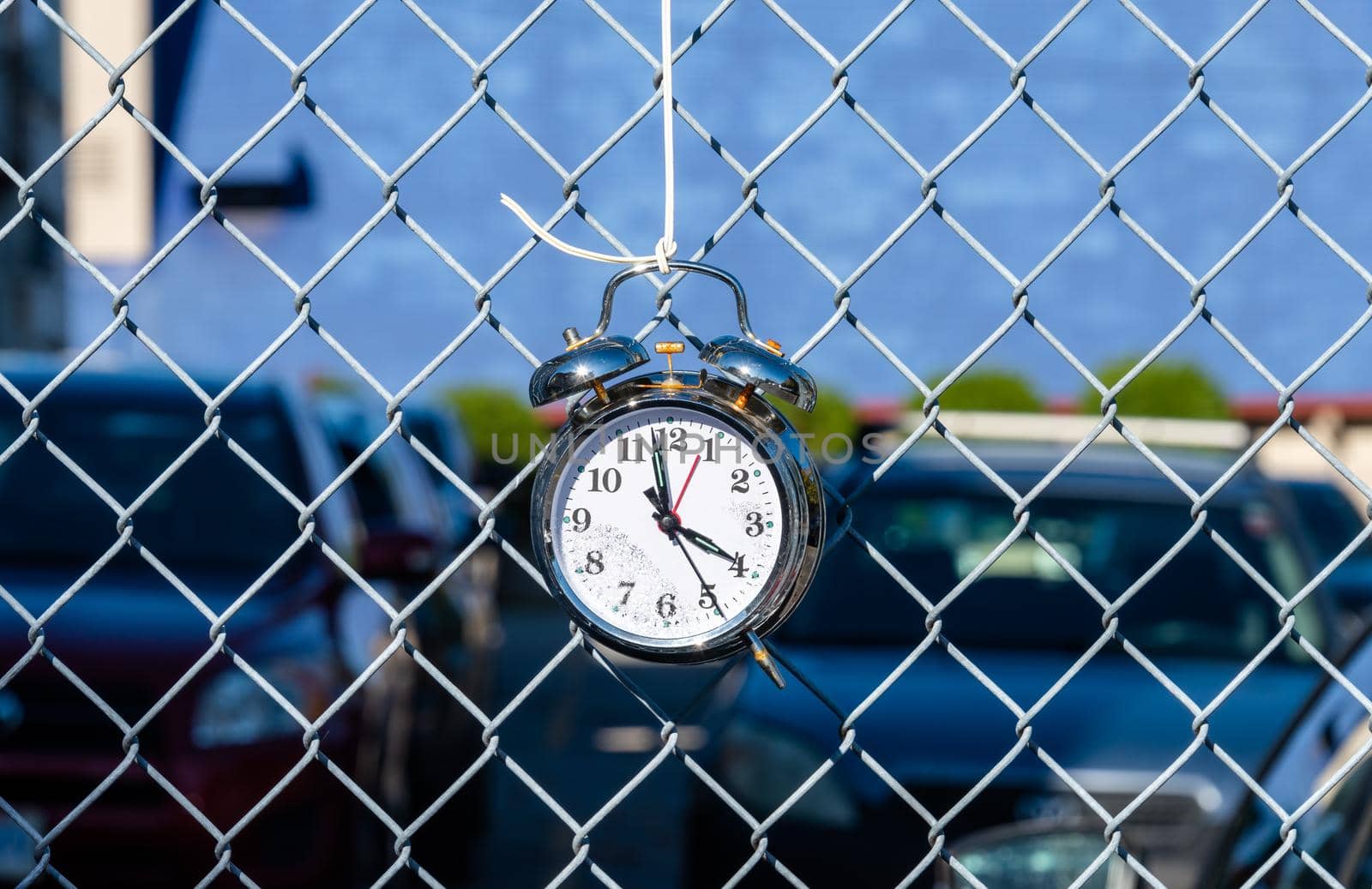Old alarm clock hanging on metal net fence with cars parked on the backgound.
