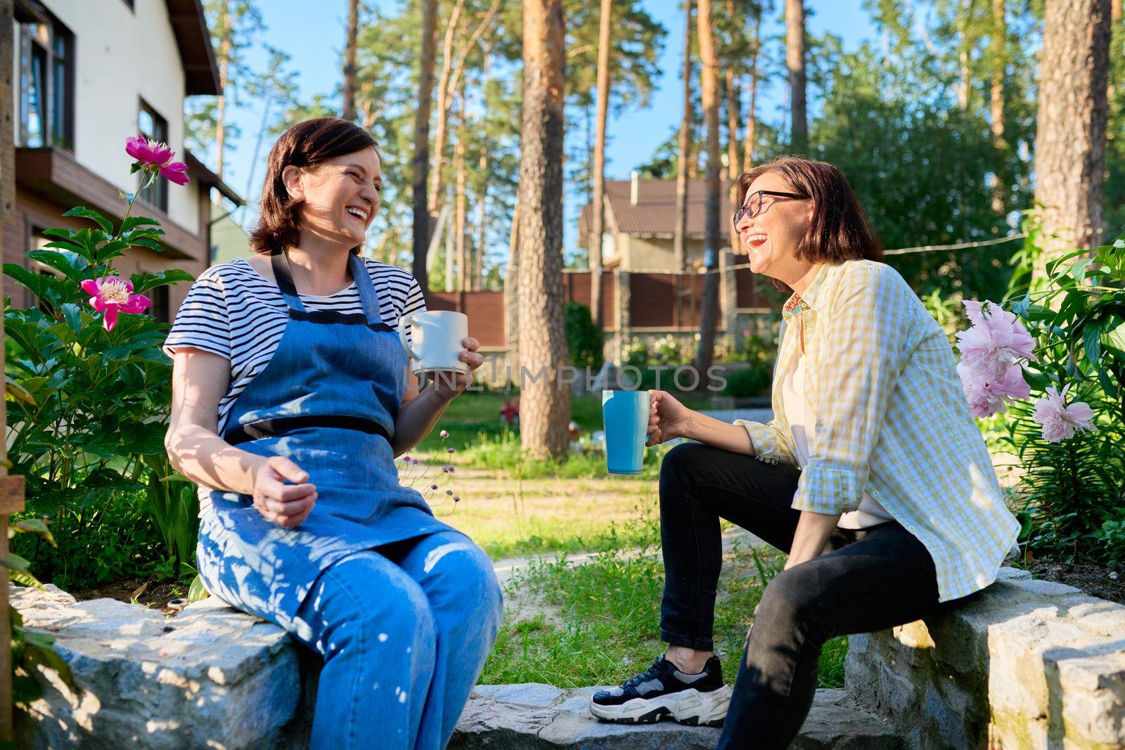 40s people, women, leisure, communication, friendship concept. Two laughing happy middle aged women sitting outdoors together in a spring garden, backyard with mugs of tea.