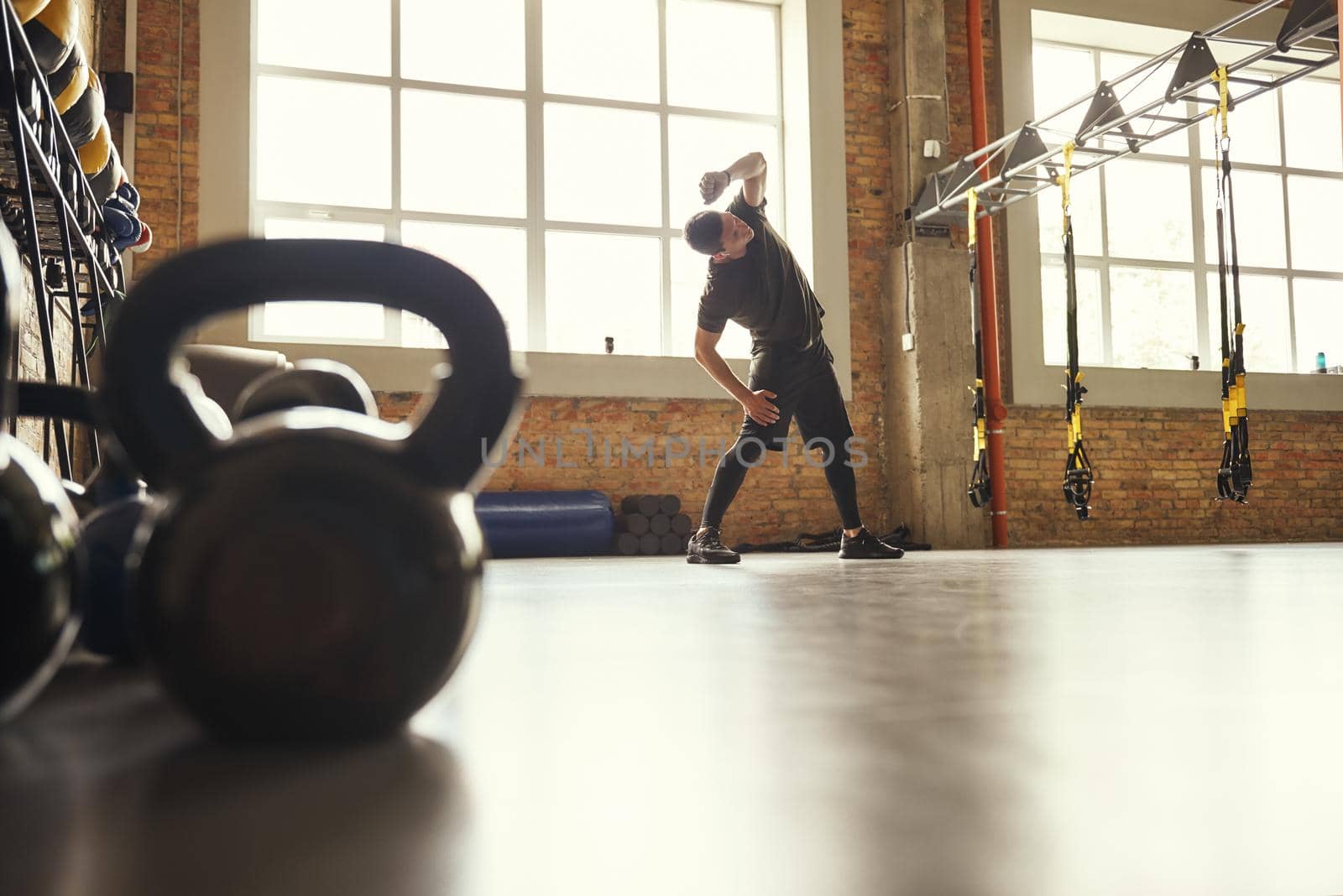 Warming up before training. Young sporty man in sportswear doing stretching exercises while standing in front of window at gym. Workout concept. Professional sport. TRX Training.