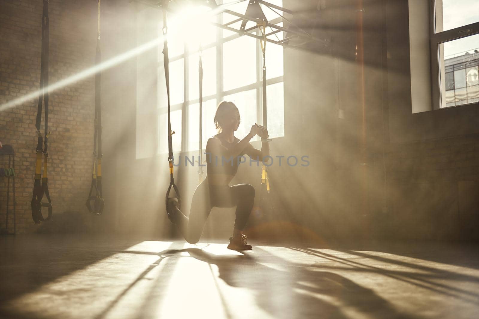Deep squat. Full-length of young athletic woman in sports clothing training legs with trx fitness straps in the gym by friendsstock