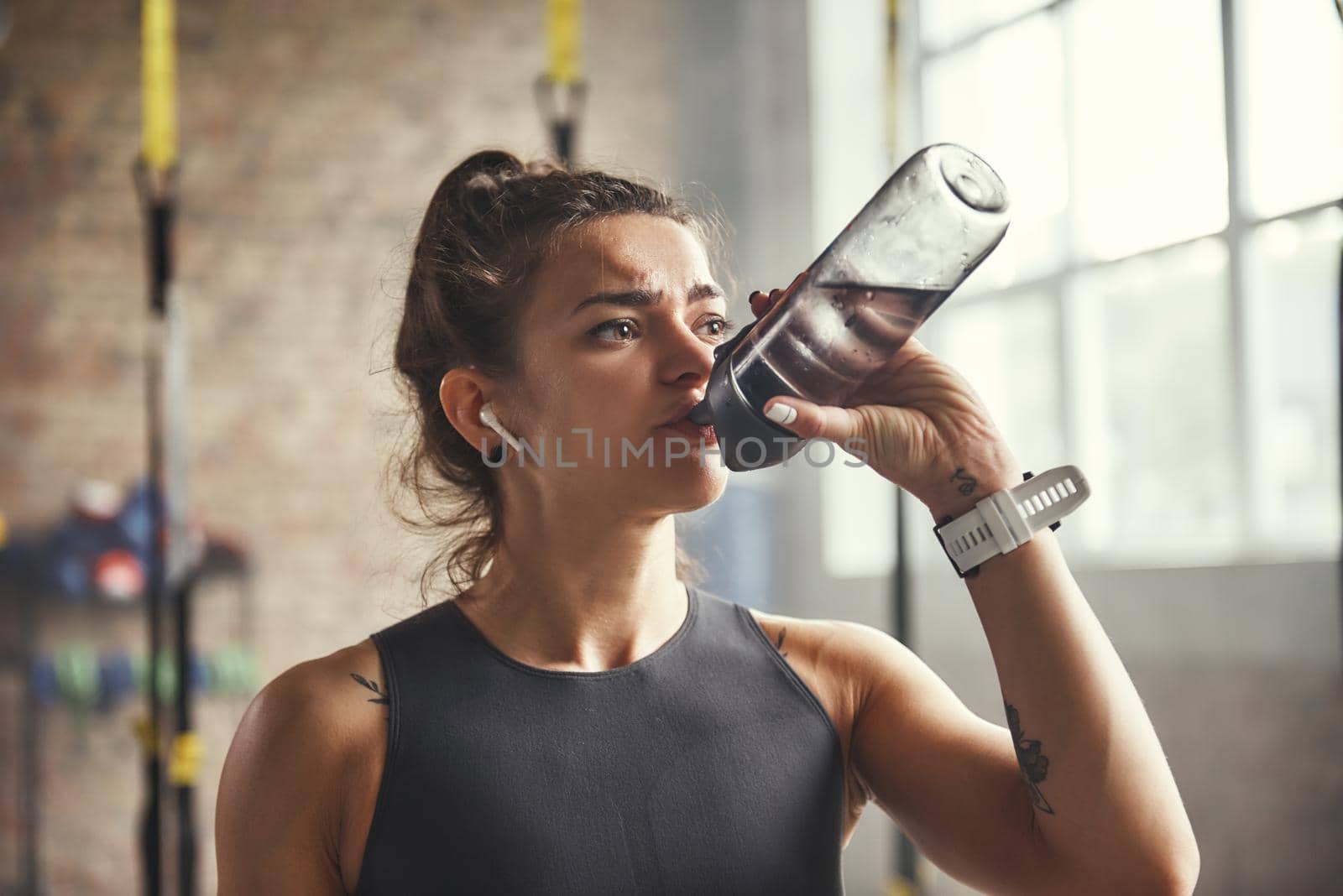 Close up of young athletic woman in earphones is drinking water while exercising in gym. Professional sport. TRX Training. Healthy lifestyle