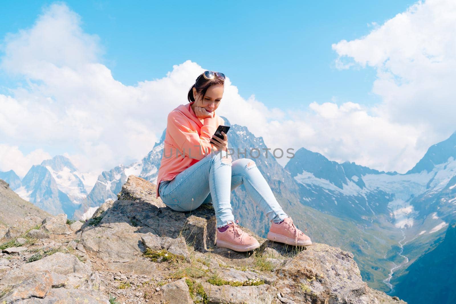 Female traveler using smartphone in mountains. Full body woman sitting on rock and browsing smartphone against cloudy sky on sunny day in mountains by epidemiks