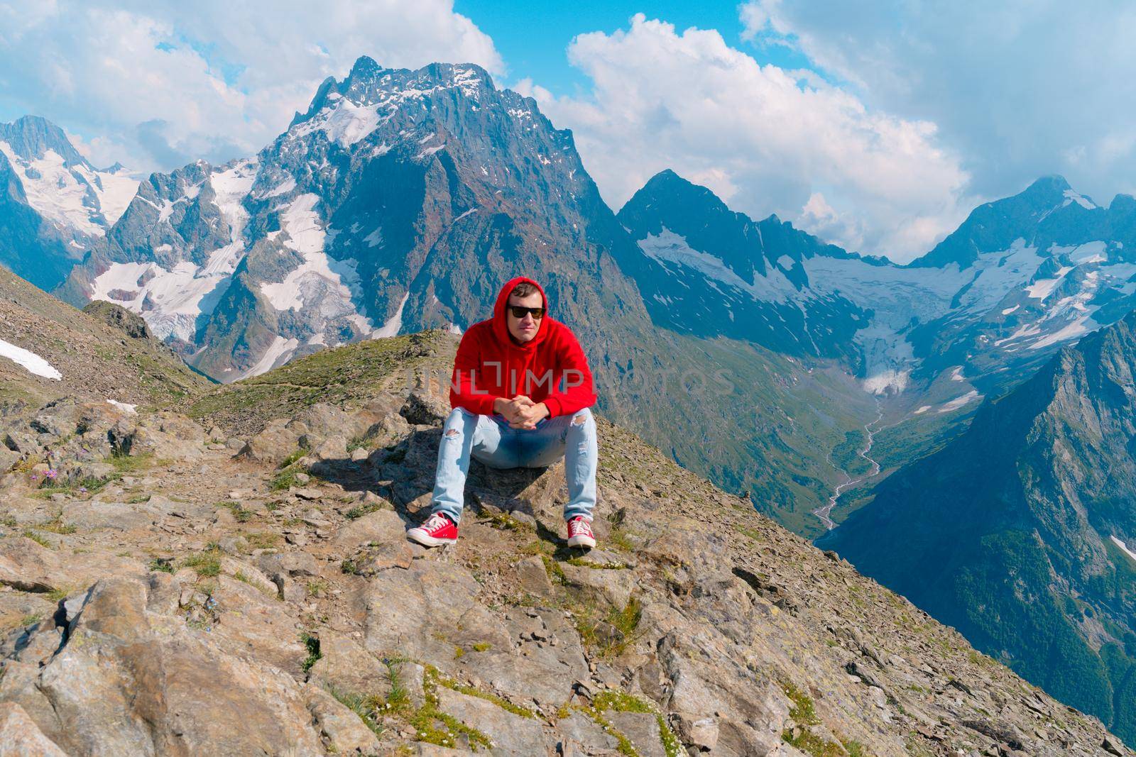 Young man in sunglasses sitting on mountain in sunny weather. Adult male in red hoodie with hood enjoying beautiful view in mountainous area. by epidemiks
