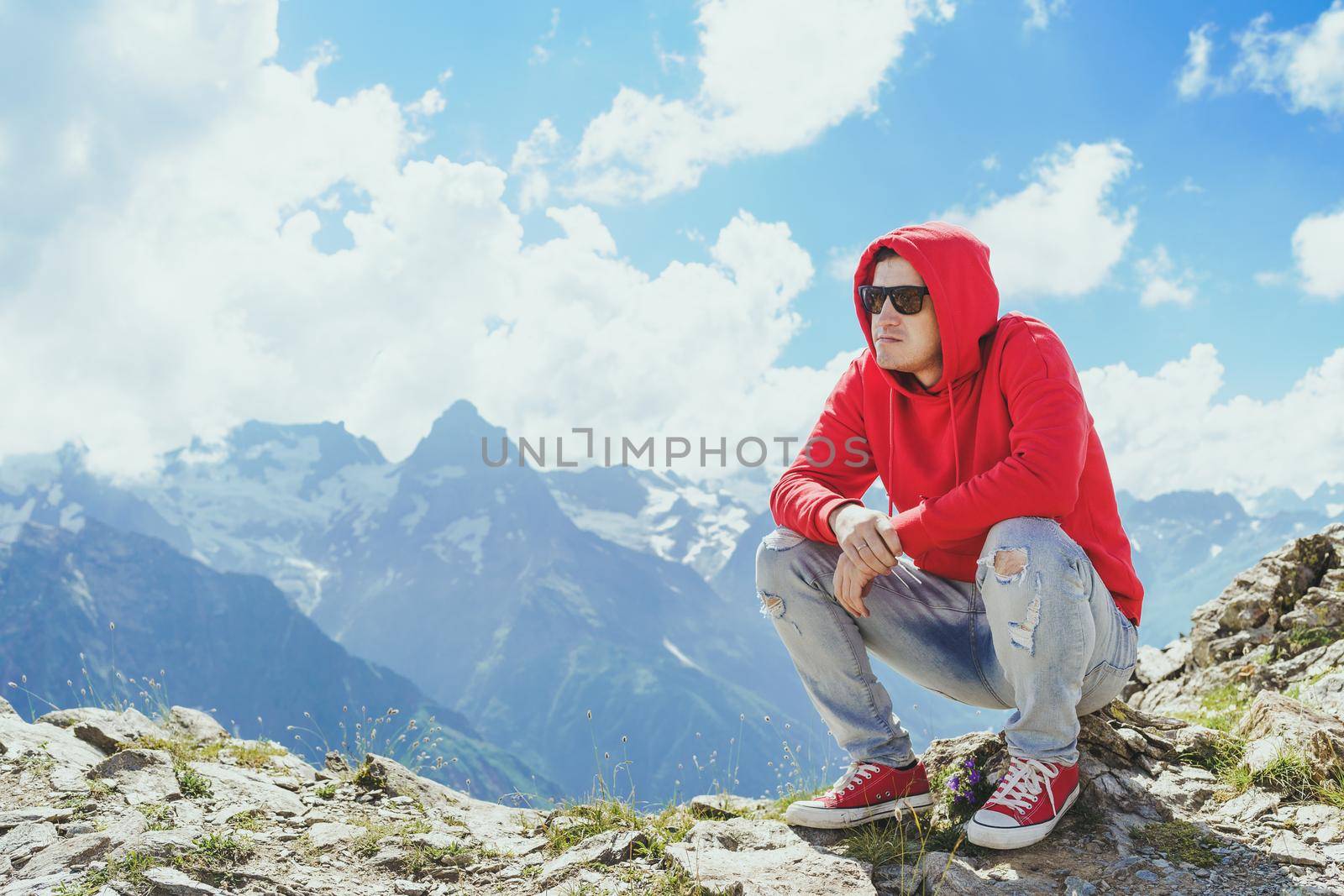 Young man in sunglasses sitting on mountain in sunny weather. Adult male in red hoodie with hood enjoying beautiful view in mountainous area. by epidemiks