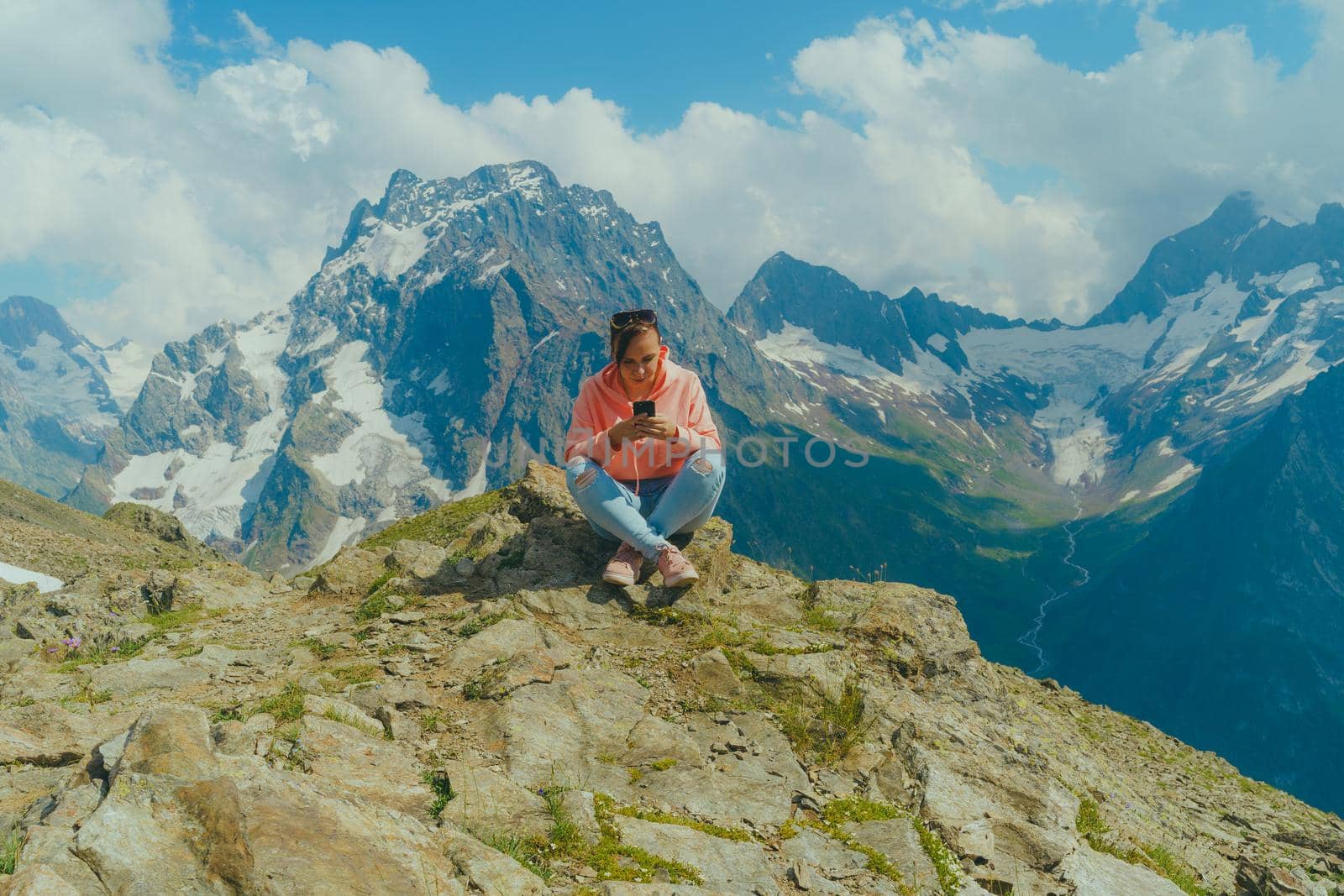 Female traveler using smartphone in mountains. Full body woman sitting on rock and browsing smartphone against cloudy sky on sunny day in mountains by epidemiks