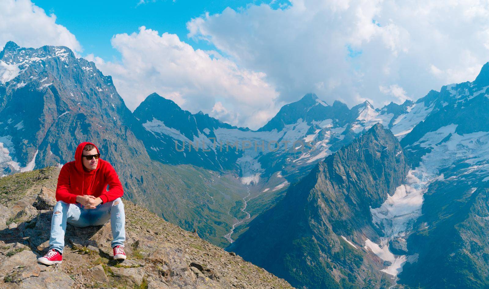 Young man in sunglasses sitting on mountain in sunny weather. Adult male in red hoodie with hood enjoying beautiful view in mountainous area. by epidemiks