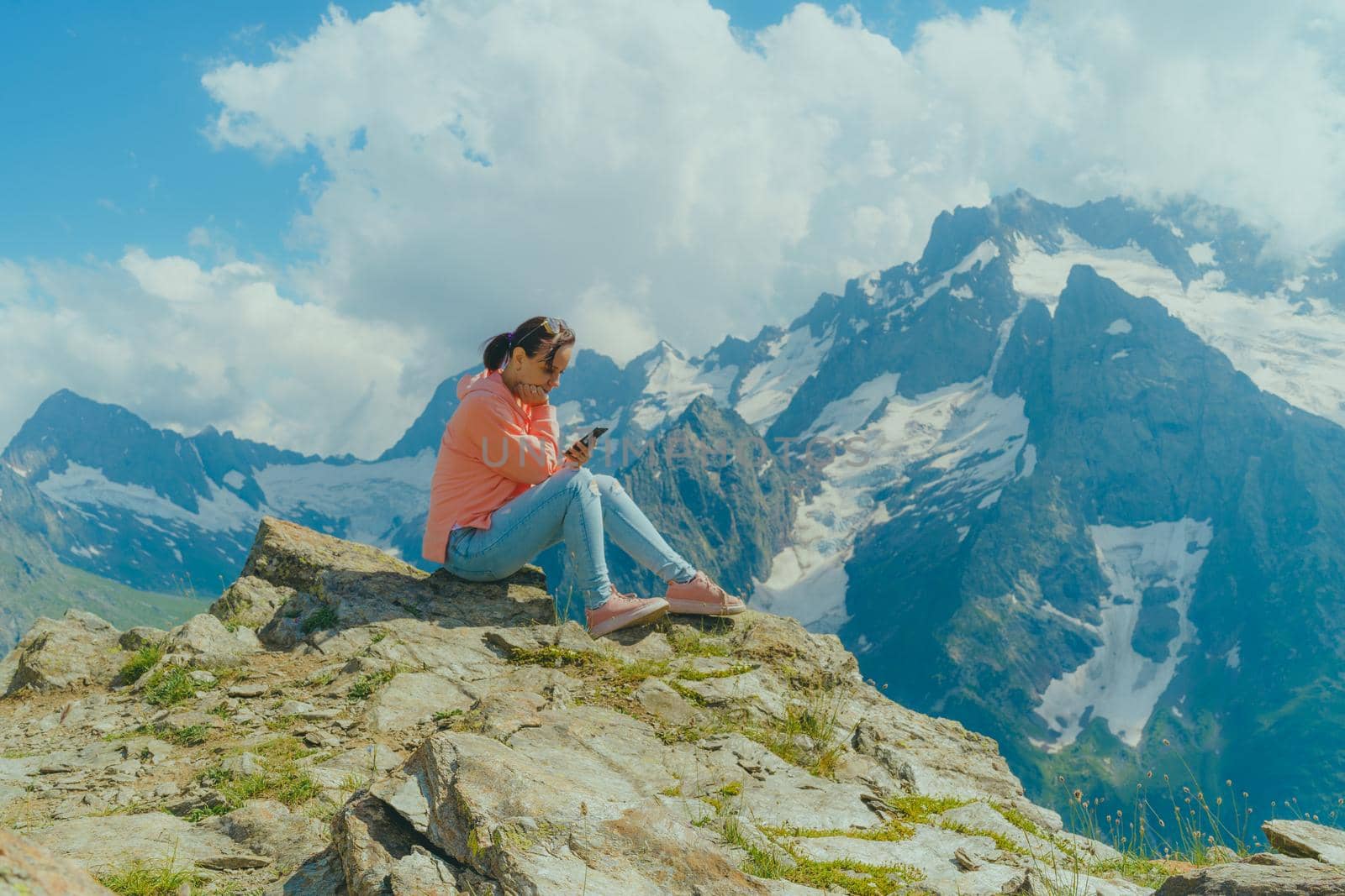 Female traveler using smartphone in mountains. Full body woman sitting on rock and browsing smartphone against cloudy sky on sunny day in mountains by epidemiks