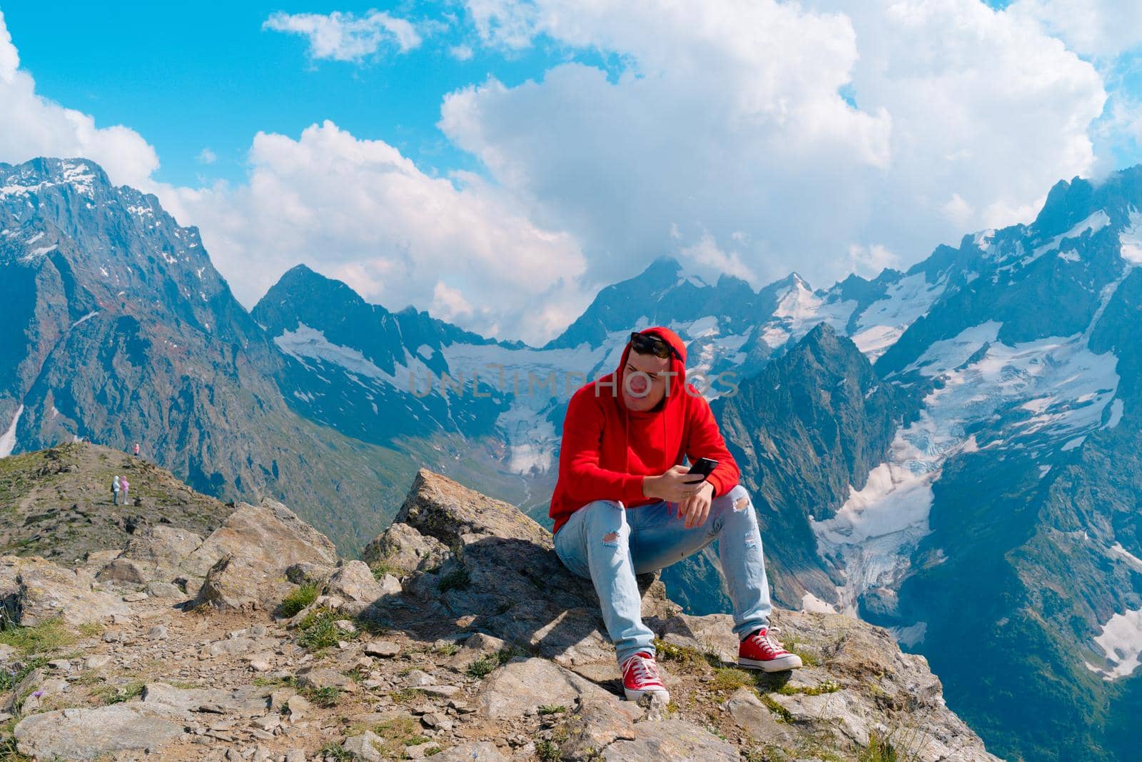 Man tourist sitting on rock with smartphone in mountains. Male traveler using mobile phone against cloudy sky on sunny day in mountainous terrain. by epidemiks