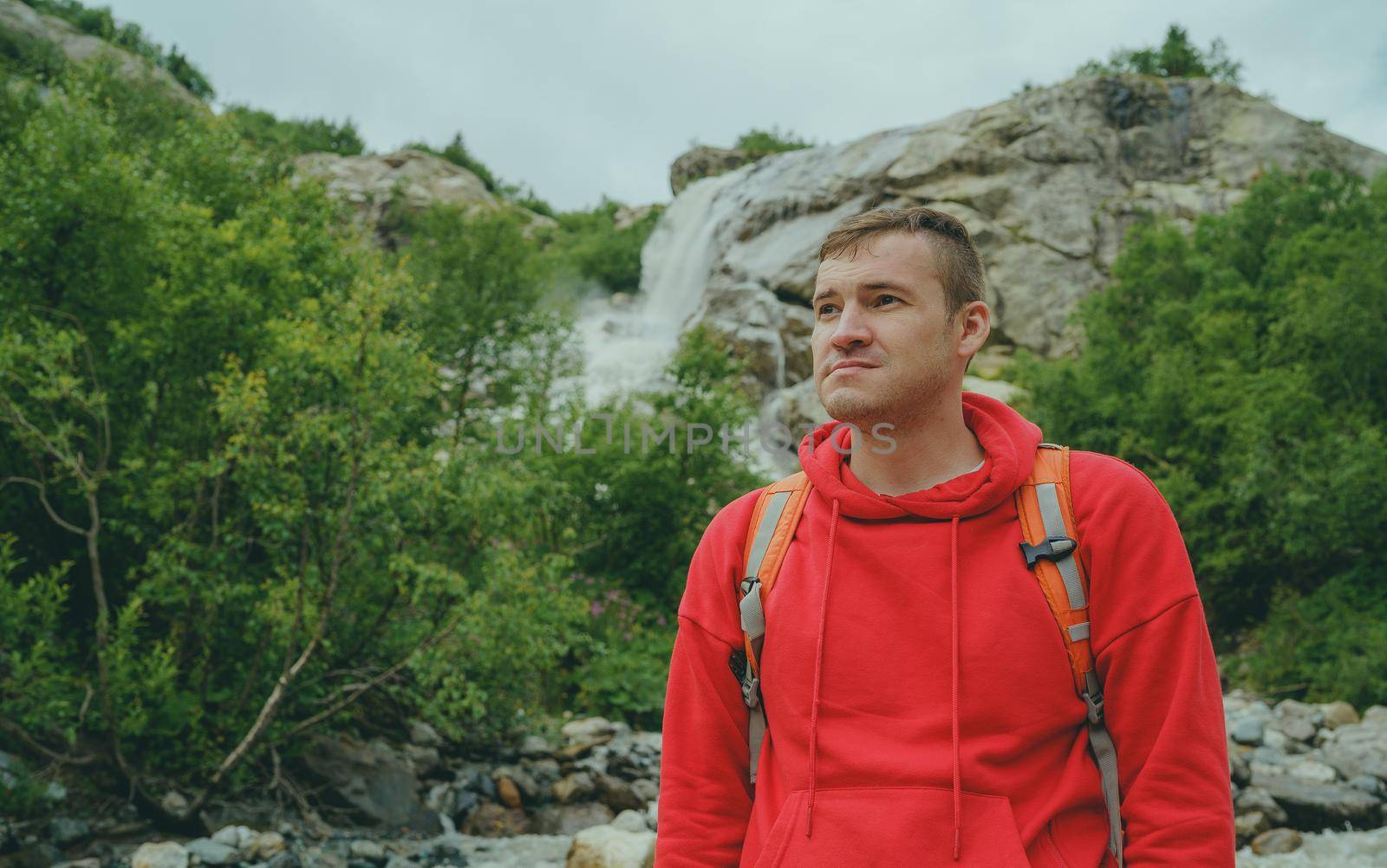 Portrait of young man looking away on background of waterfall. Adult male tourist enjoying beautiful view in mountainous area. by epidemiks