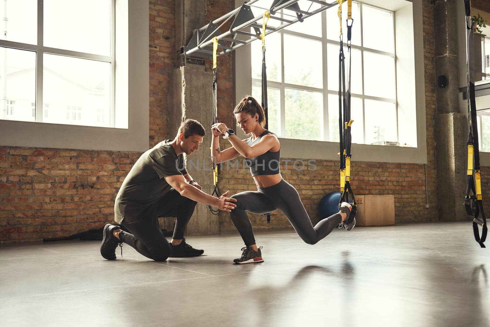 Doing squat exercise. Confident young personal trainer is showing slim athletic woman how to do squats with Trx fitness straps while training at gym. by friendsstock