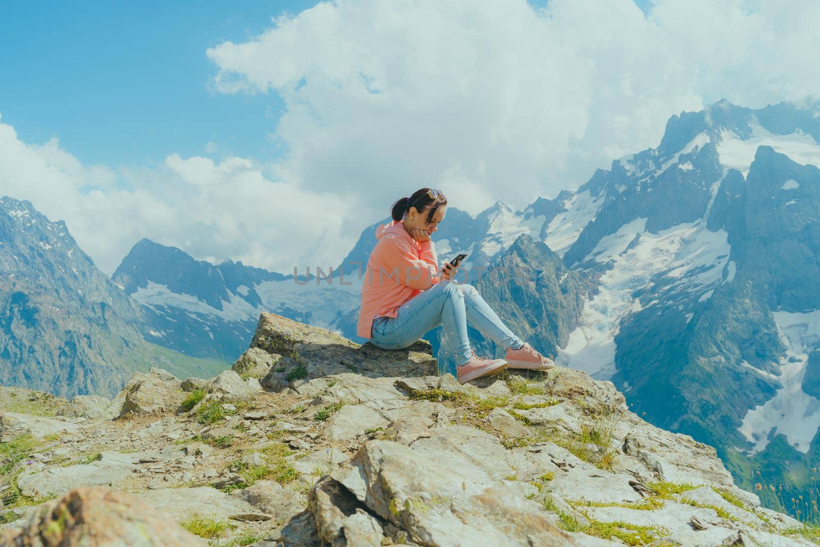 Full body woman sitting on rock and browsing smartphone against cloudy sky on sunny day in mountains