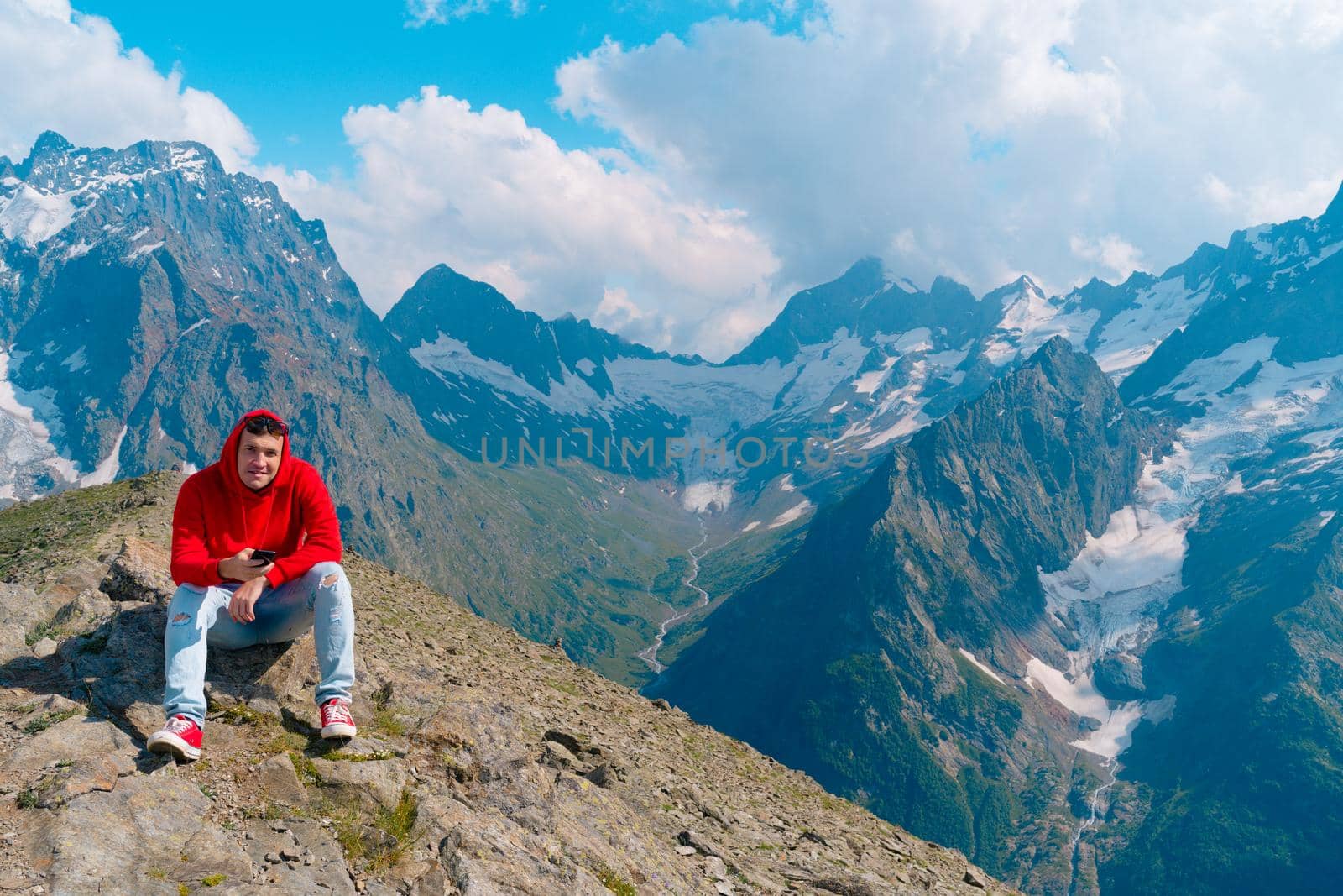 Man tourist sitting on rock with smartphone in mountains. Male traveler using mobile phone against cloudy sky on sunny day in mountainous terrain. by epidemiks