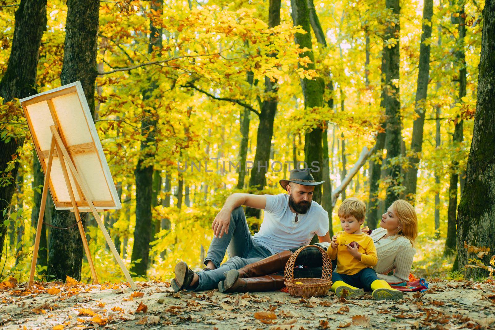 Young family of artists doing a picnic on an autumns day. The concept of a happy family. Young smiling family doing a picnic on an autumns day. Drawing easel with artwork. Inspiration. by Tverdokhlib