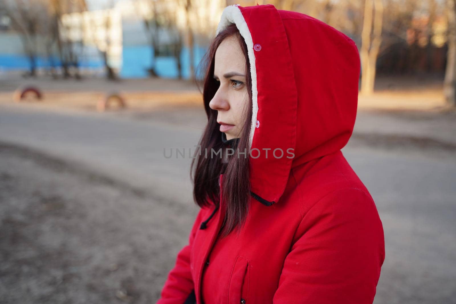 Portrait of young woman in red hood and jacket on street. Adult serious female looks away thoughtfully, standing on fresh air. by epidemiks