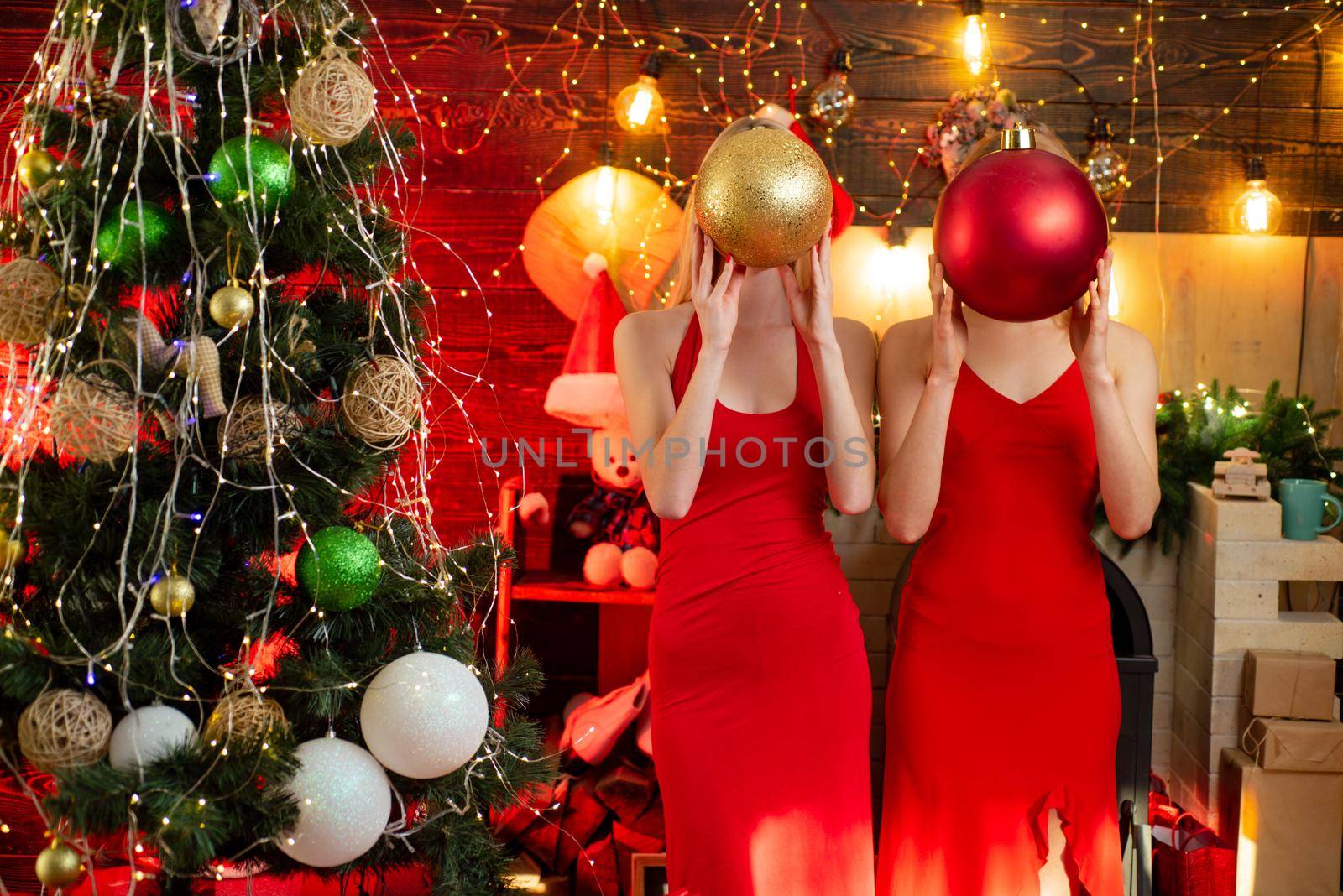 Two sexy hot girls in red dresses hide their faces with Christmas tree decorating balls at beautiful winter home background. Xmas tree. Concept of Merry Christmas and New Year