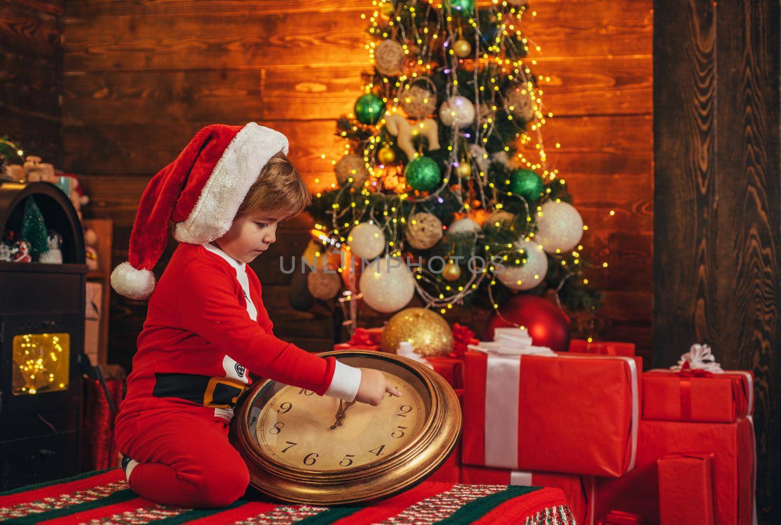 Little Santa kid waiting a New Year midnight and holding clock. Little genius. Gifts for winter holidays at fire place. Santa boy celebrate christmas at home. Gift card concept
