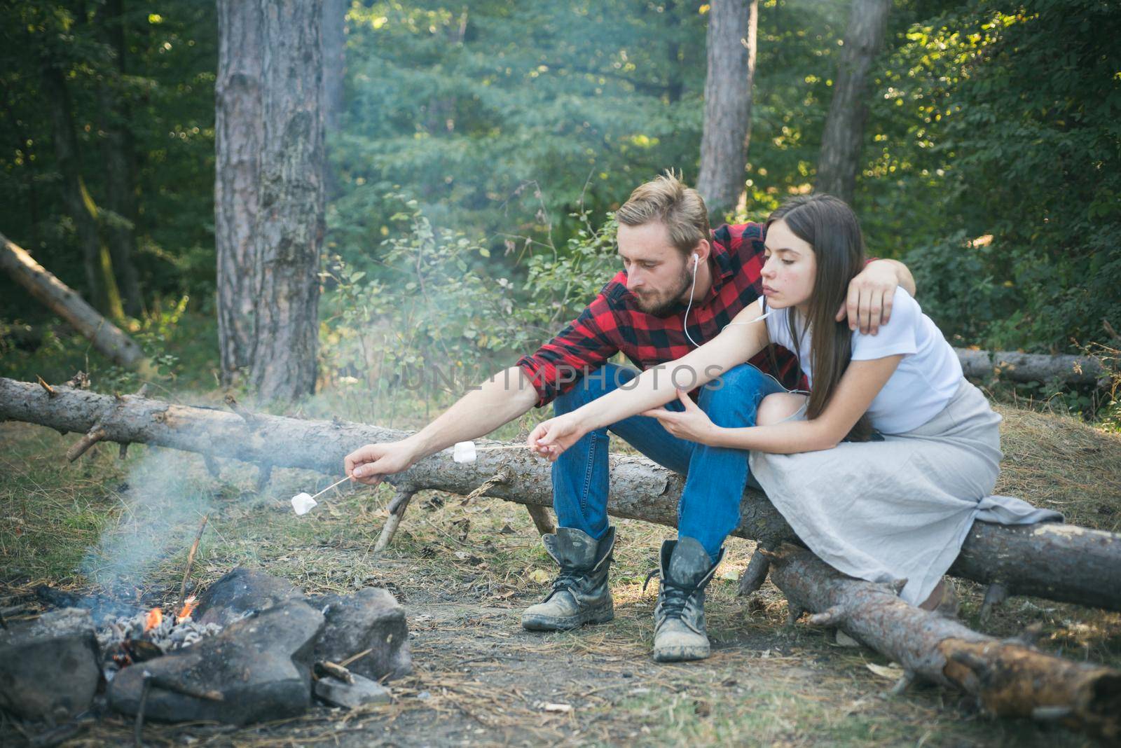 Pretty couple roast marshmallow candies on the campfire in forest. Tourists relaxing. Picnic time. Tourism concept. Young couple having picnic in woods. Camp fire in summer