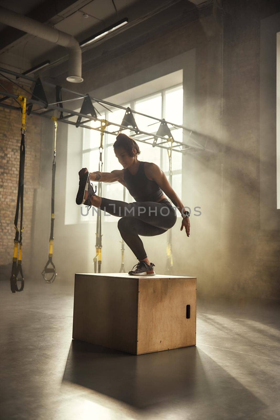 Becoming the best. Side view of athletic woman in sportswear doing squat and training legs while standing on wooden box at gym. by friendsstock