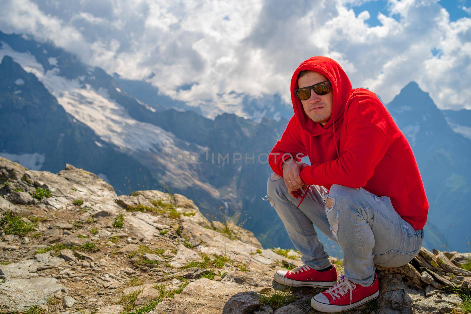 Young man in sunglasses sitting on mountain in sunny weather. Adult male in red hoodie with hood enjoying beautiful view in mountainous area. by epidemiks