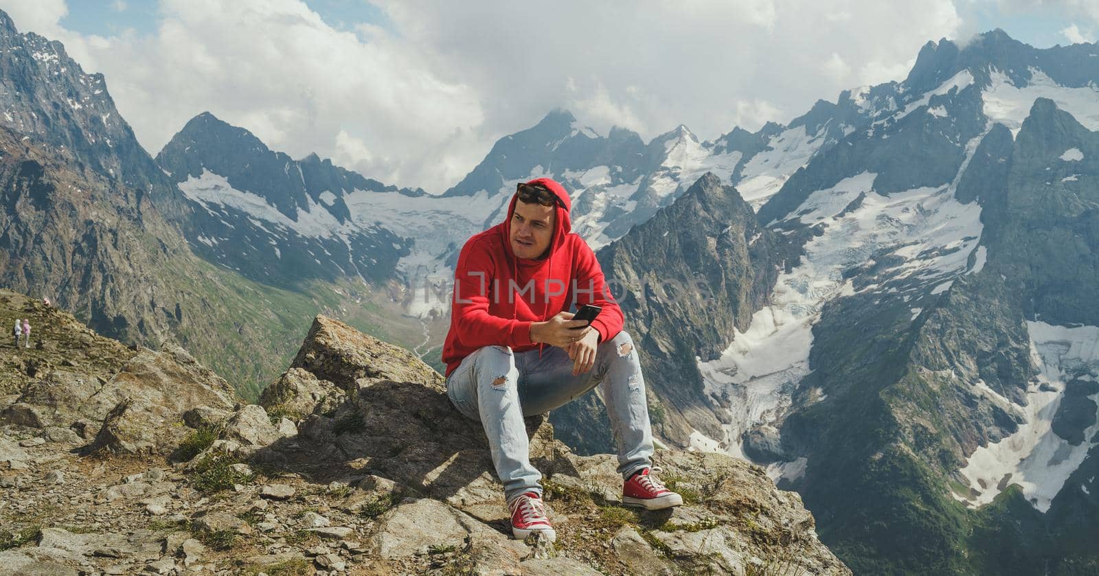 Man tourist sitting on rock with smartphone in mountains. Male traveler using mobile phone against cloudy sky on sunny day in mountainous terrain. by epidemiks